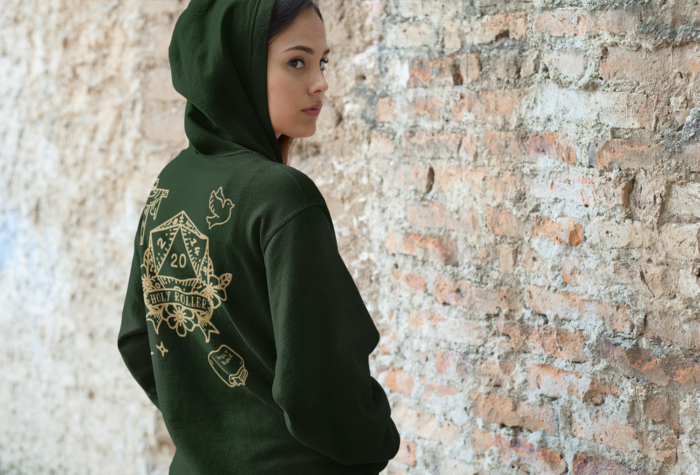 A woman wearing a dark forest green hoodie with a large gold graphic print of a DnD Die with a banner that reads" Holy Roller", on the back, standing against a rustic brick wall.
